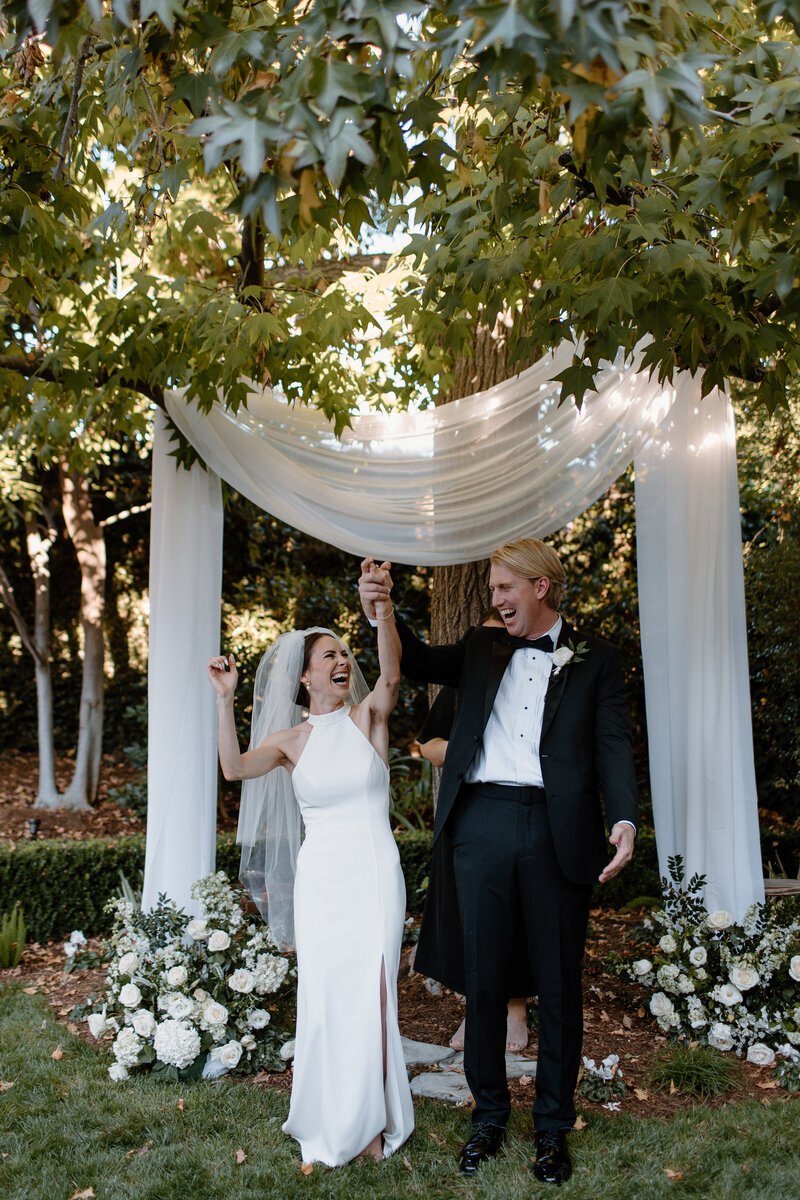 7-radiant-love-event-2021-outdoor-ceremony-bride-groom-holding-hands-up-cheering-white-flowy-draping-tree-backdrop-romantic-elegant-timeless