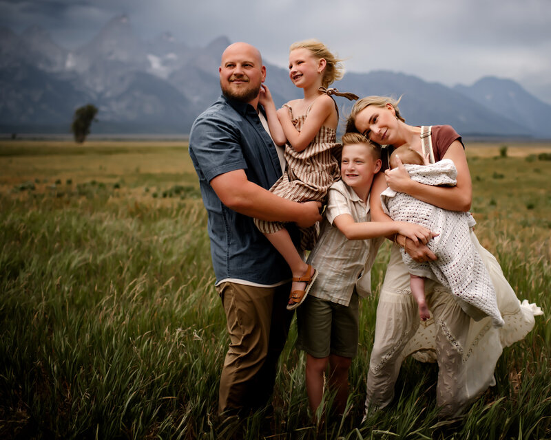A damily of five stand together in a lush green Jackson, WY field while the wind blows. 