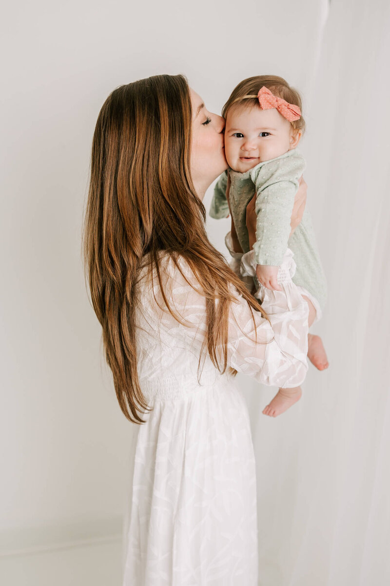 Mom wearing white dress, holding daughter during their motherhood family session in the studio.