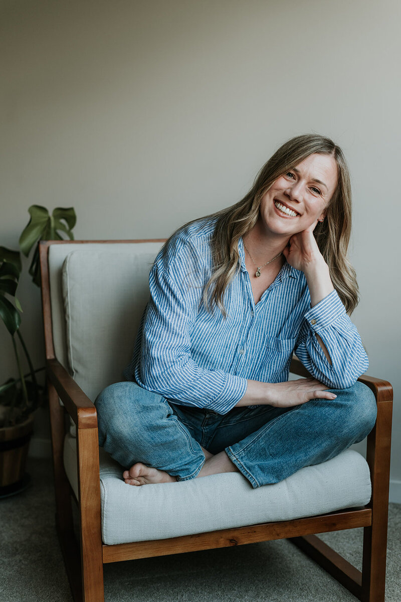 female wearing jeans and blue striped shirt sitting cross legged on wooden chair smiling with hand resting on face