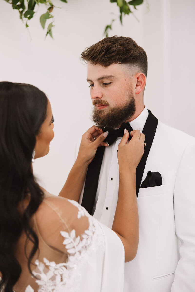 Bride adjusting groom’s black bow tie as he wears a white tuxedo with black lapels and pocket square, captured in a romantic pre-ceremony moment by Wells Design Co
