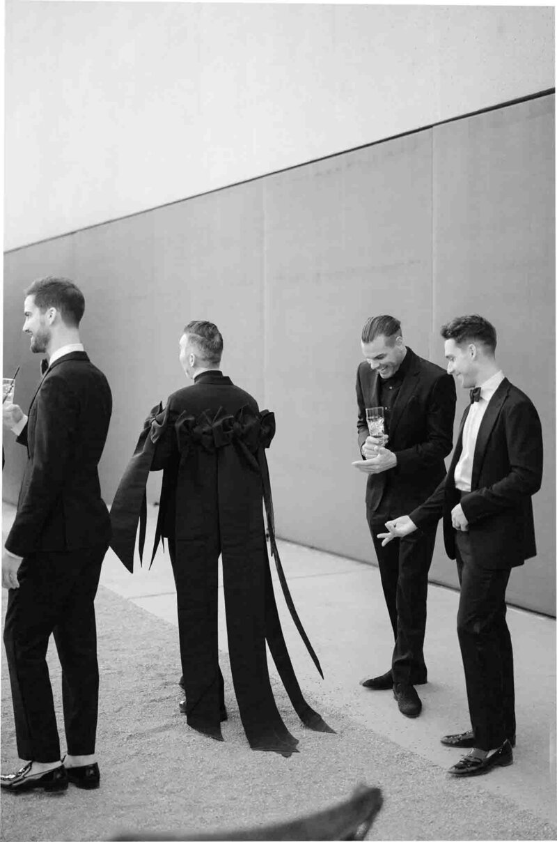 black and white photo of two wedding gusts admiring another guest's fashion forward black suit adorned with large bows