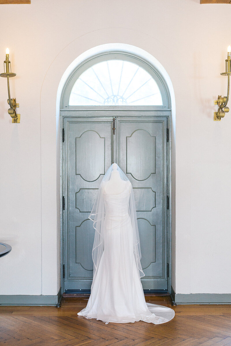 White wedding dress and veil hang on mannequin in front of blue doors inside Crane Cottage