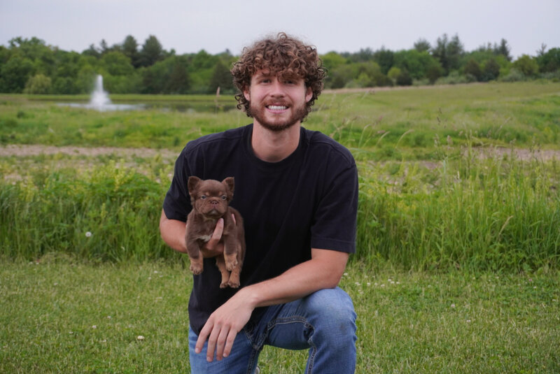 Myles Clawson owners of Pawz N Clawz Puppies standing  in front of a white wall wearing a white shirt with his business logo smiling.