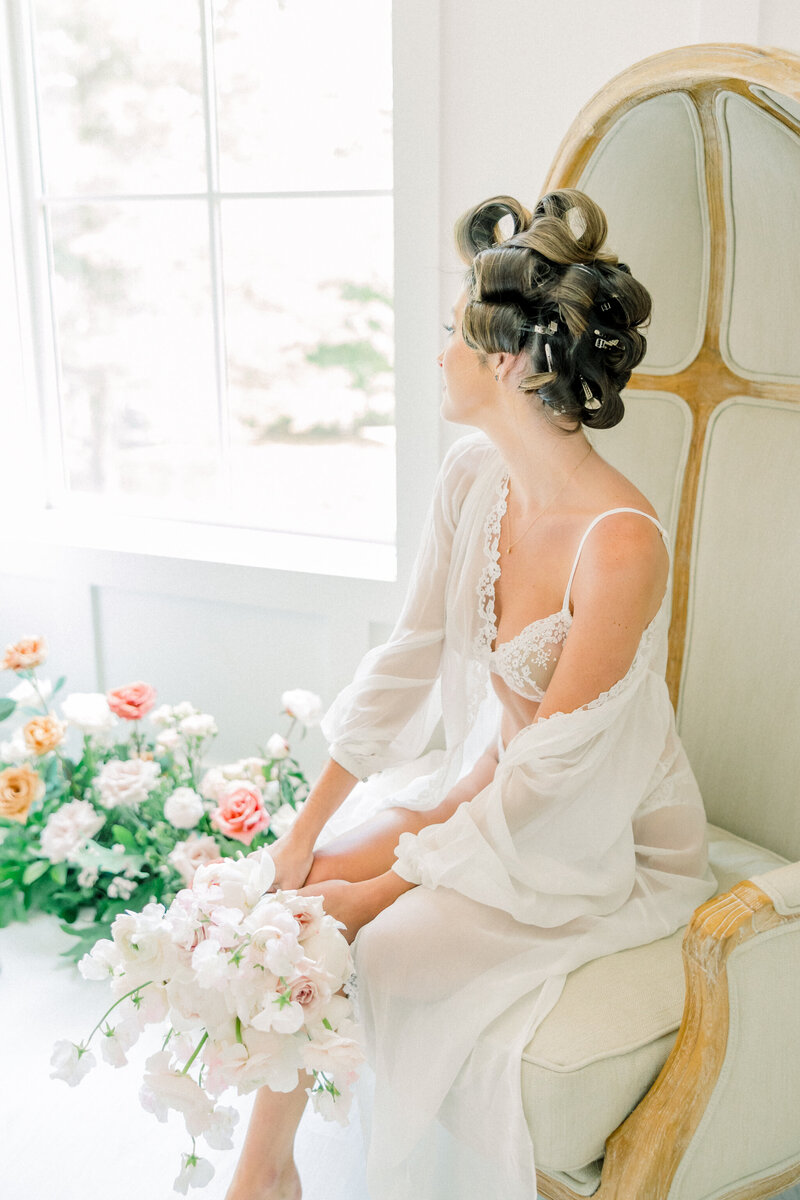 Richmond VA Boudoir Photographer | A girl in white floral lace lingerie and sheer white robe draped off her shoulder sits in a chair looking out the window. She holds a delicate, romantic bouquet of white and soft pink florals.
