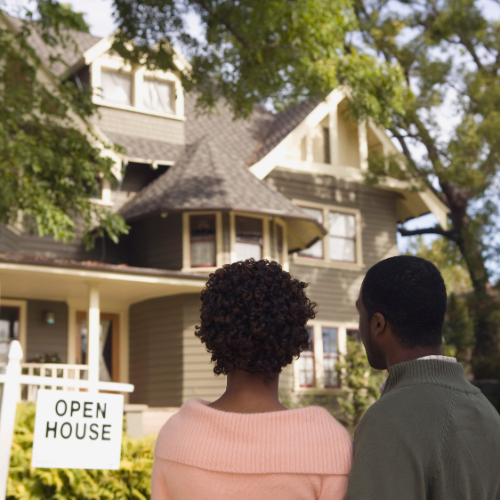 “A young Black couple stands together outside a Victorian-style home with an ‘Open House’ sign on the lawn. They are looking toward the house, symbolizing the dream of homeownership and new beginnings.