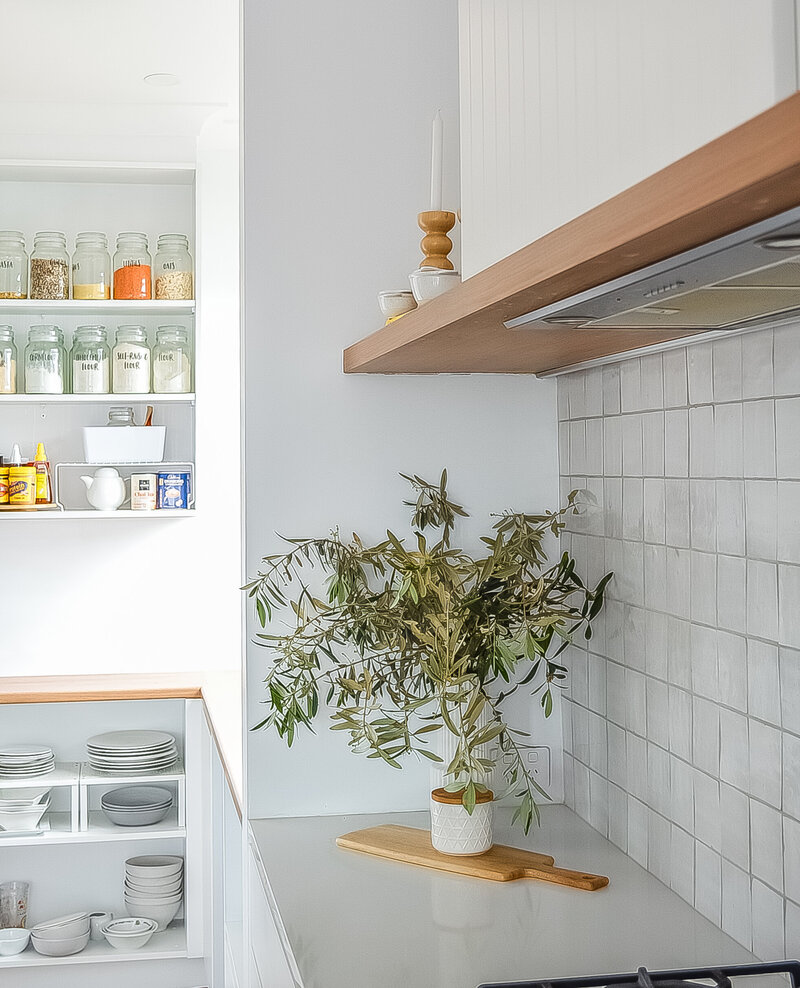 A minimalist kitchen reflects bespoke interior design Newcastle NSW, featuring open shelves, jars, dishes, and a potted plant on a pristine white countertop.