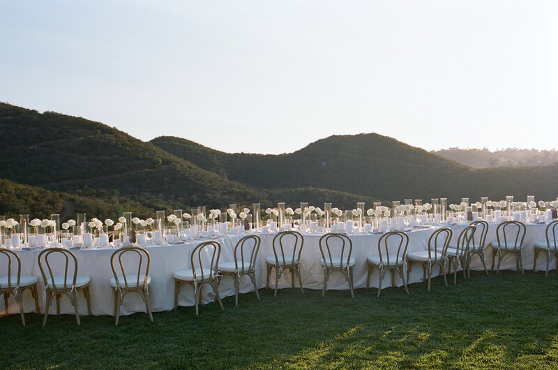 BP Collective European Inspired Experience with a long alfresco dining table set with white florals and candles against rolling hillside views.