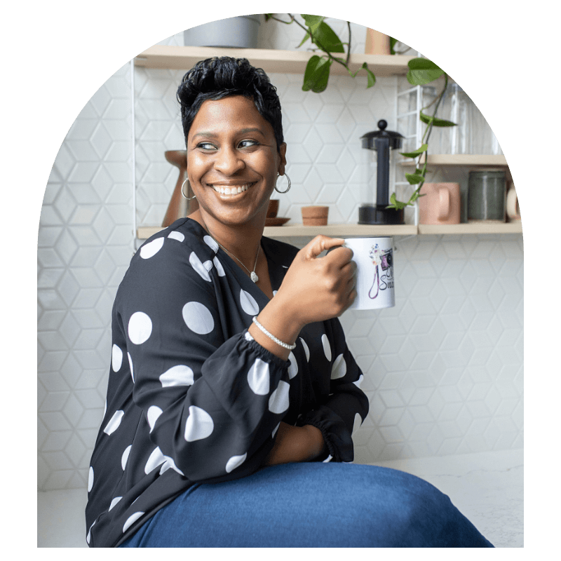 Metro Detroit Portrait Photographer  with short pixie haircut in black polka dot blouse and blue jean jeggings sitting on counter with coffee mug posing causually