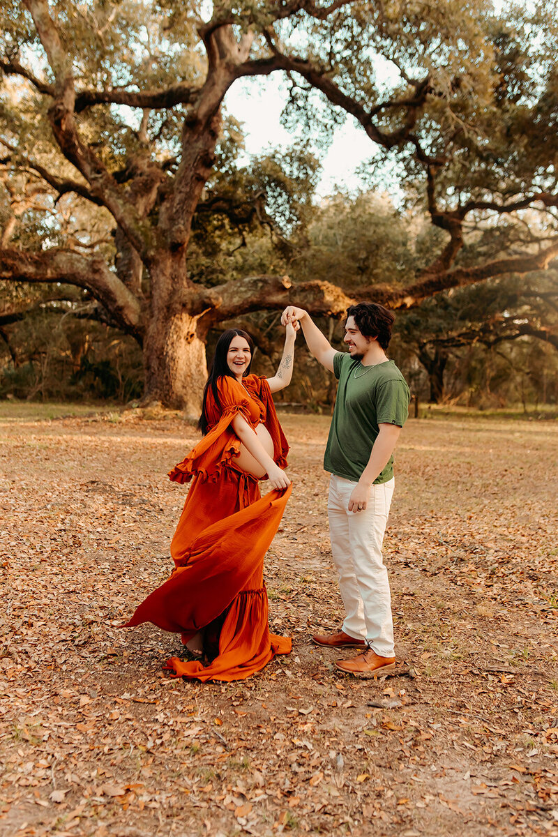 Maternity & couples photography session in Cleveland, Texas, featuring a man twirling a pregnant woman in an orange dress beneath oak trees