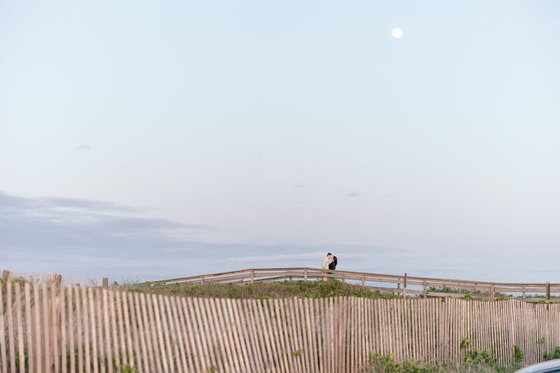 Engaged couple embracing on Duxbury Beach near Marshfield, with the moon rising above a scenic bridge at sunset.
