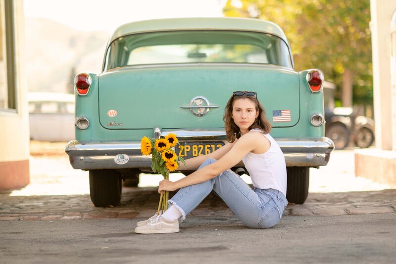 High school senior girl wearing a white tank top and blue jeans, holding sunflowers while sitting in front of a teal car