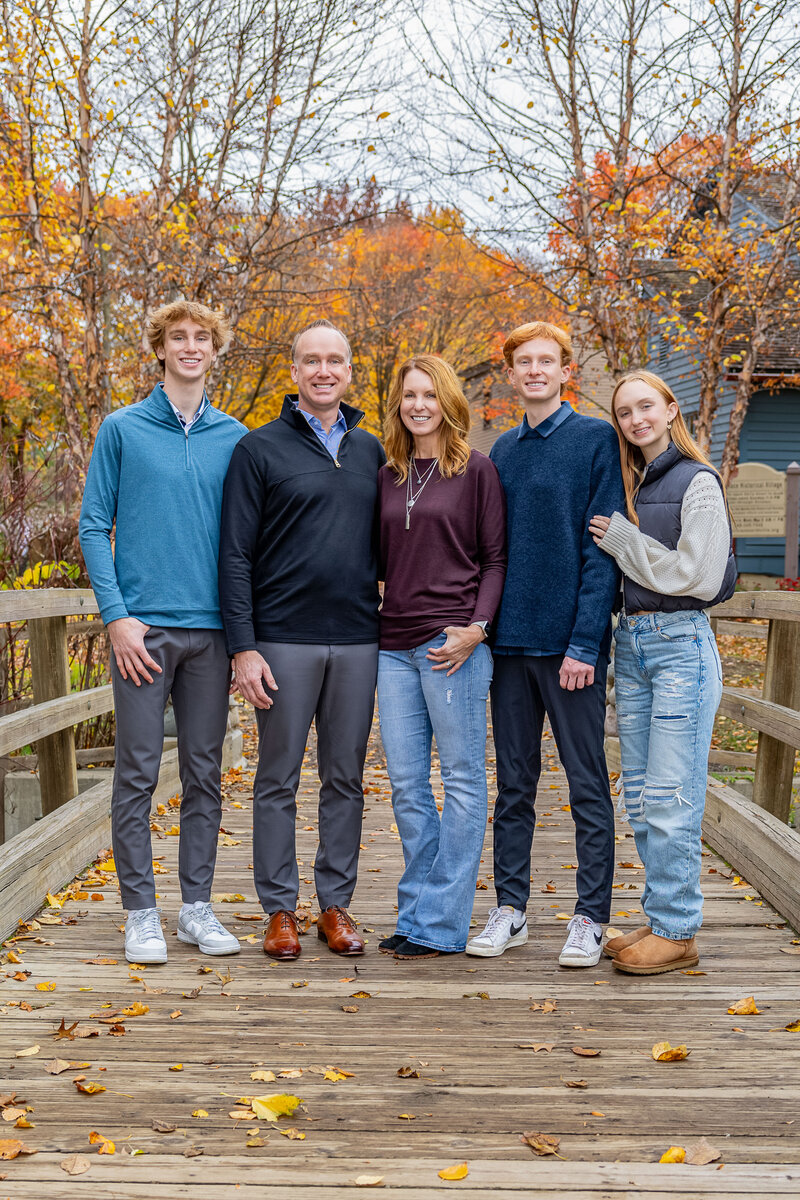 A family of 5 is standing together on a bridge for a portrait.  There are orange and yellow colored trees in the distance