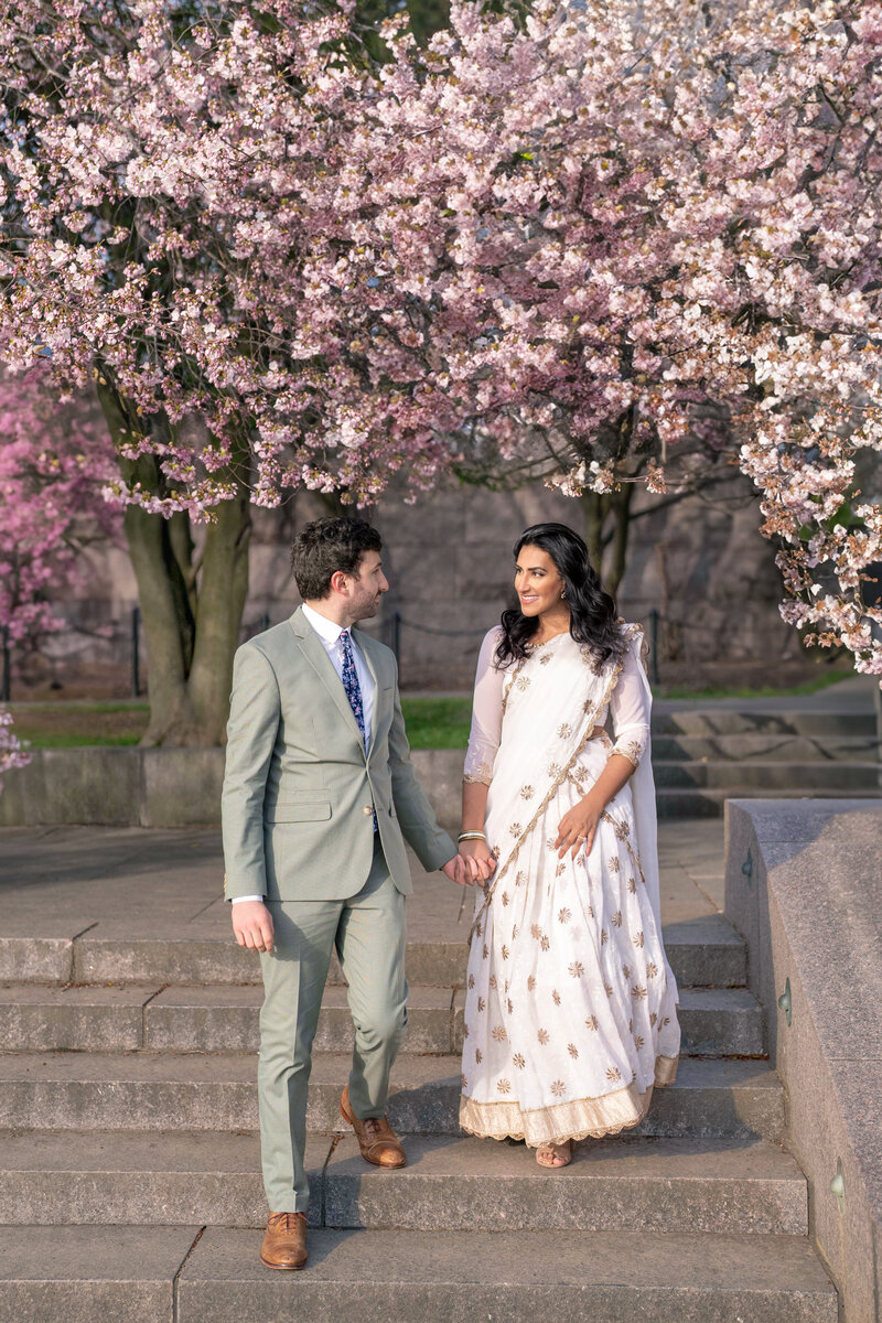 A couple walks hand in hand down stone steps surrounded by blooming cherry blossom trees. The man wears a light gray suit, and the woman is in a white and gold saree. They both smile at each other, enjoying the beautiful spring setting.