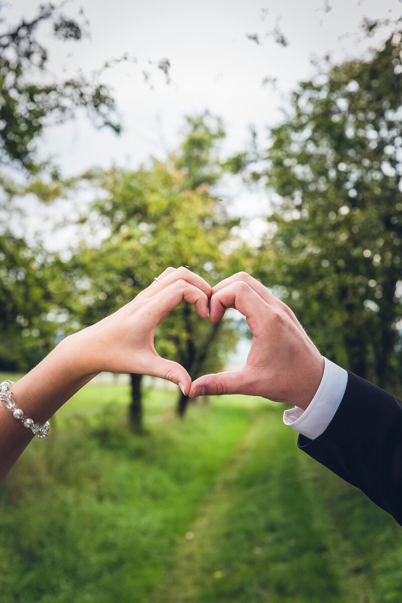 Close-up of a bride and groom forming a heart shape with their hands in front of a tree-lined path in a grassy outdoor setting.