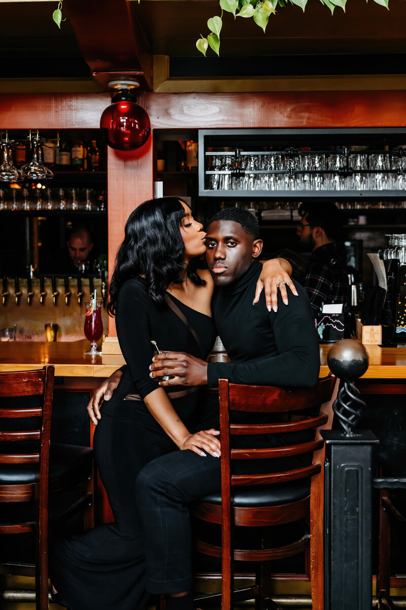a lovely BIPOC couple in front of a bar cuddling into each other