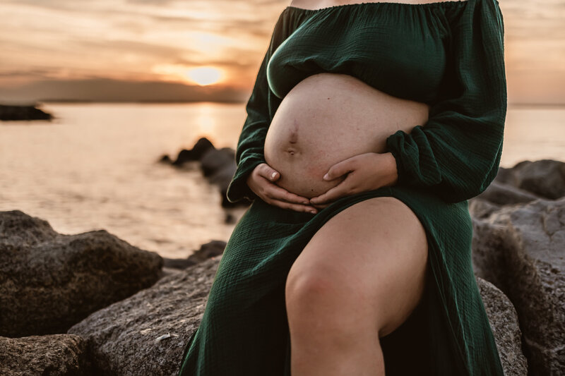 pregnant women sitting on some rocks at the beach with a beautiful sunset behind her