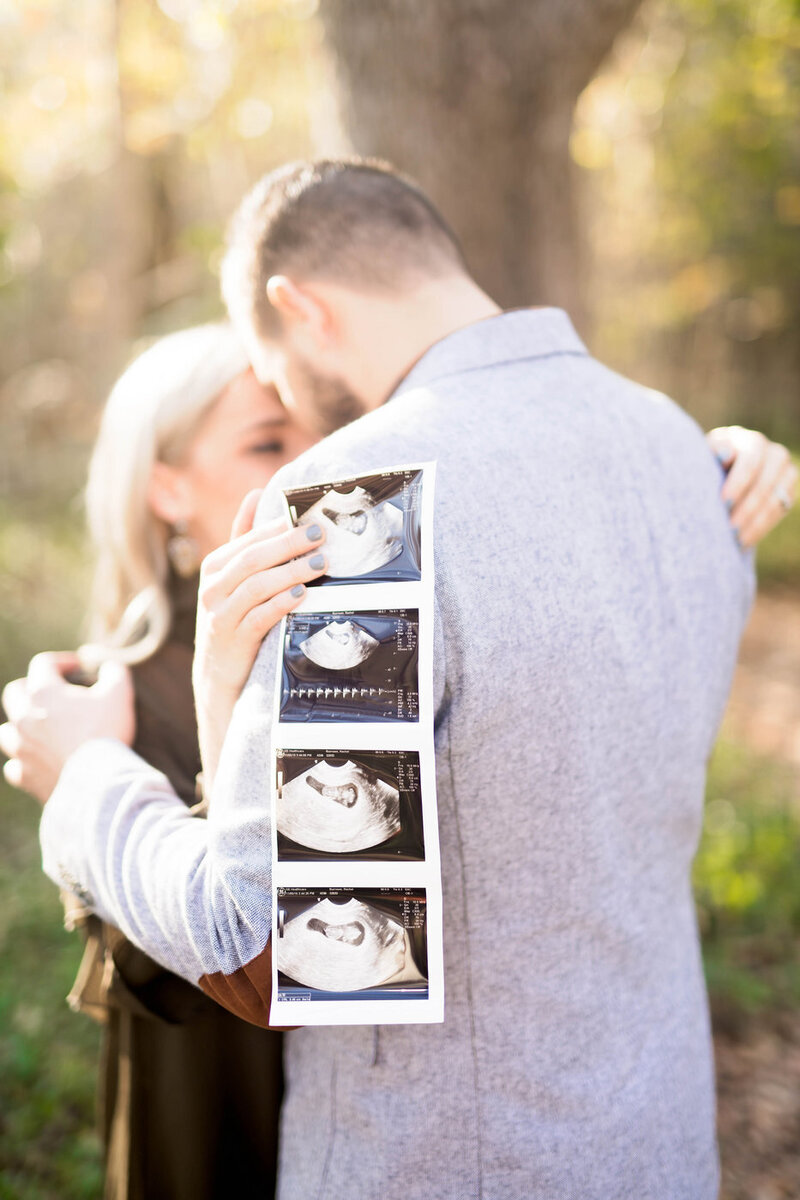 A couple is turned with their backs to the camera holding each other. The woman holds 4 ultrasound pictures over the man's shoulder.