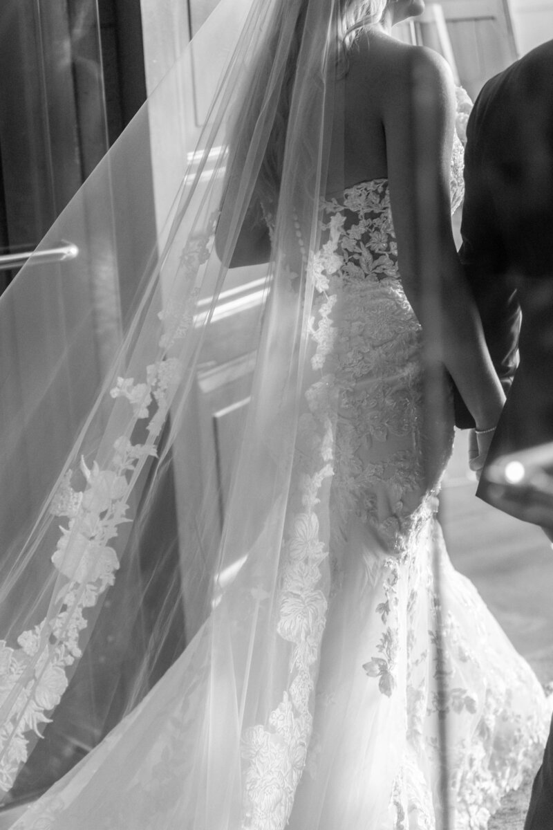 Black and white image of a bride in a lace wedding dress with a long veil walking hand in hand with a suited person. Veil and dress detail prominently visible, capturing an elegant and intimate moment.