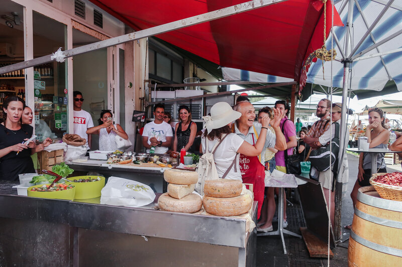 A sandwich maker dances with a customer while enthusiastic patrons look on at an outdoor sandwich shop in Italy