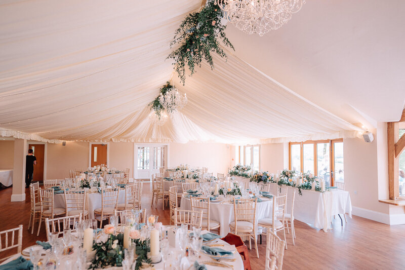 A tall gold stand of flowers with white hydrangeas, pink roses, white stocks and white peonies. Hanging jasmine trails with gold tea lights and dinner candles nestled at the base for this Cornwall wedding