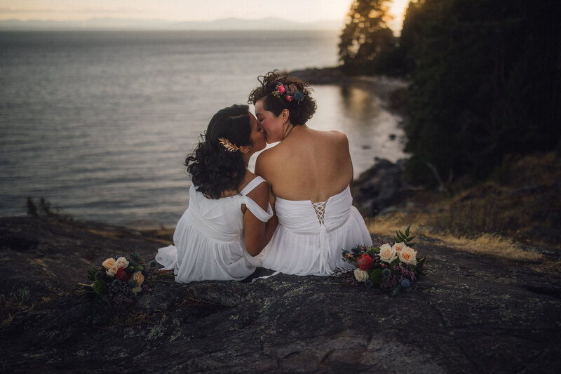 lesbian couple kissing in wedding gowns by the water at sunset