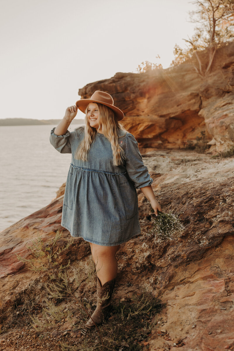 A blonde senior girl in a denim dress and cowboy boots standing by the lake.