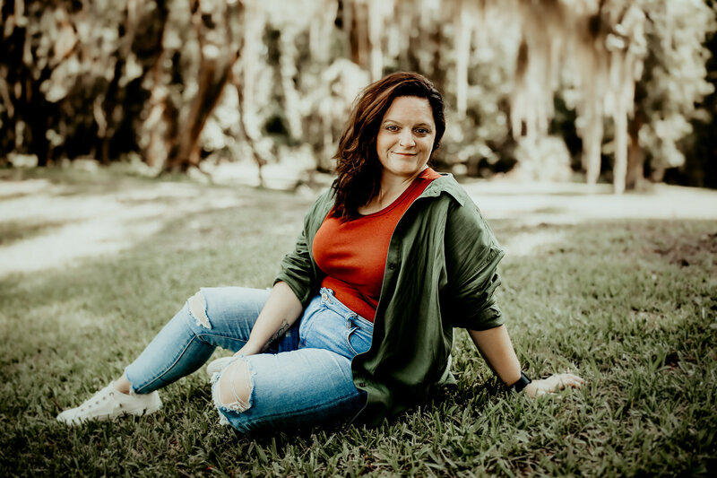 A photographer in  Macon, GA captures a woman sitting on the grass, resting her arm behind her. She wears a green jacket over a red top and ripped jeans, with trees visible in the background.