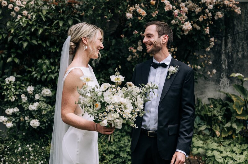 a bride holding a white green and yellow wild bouquet and a groom in a tuxedo with black bowtie in christchurch