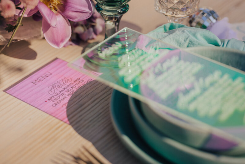 A close-up of a modern tablescape featuring a holographic acrylic menu atop teal plates, soft linens, and fresh pink florals. Disco balls, iridescent reflections, and vintage glassware add a funky-glam twist to this colorful setup. Captured by Dani Lacey Photographs.