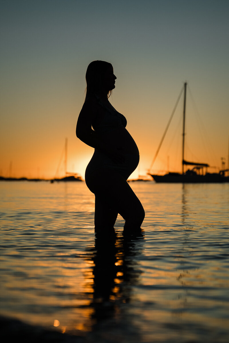pregnant lady in water in front of a sunset and some boats