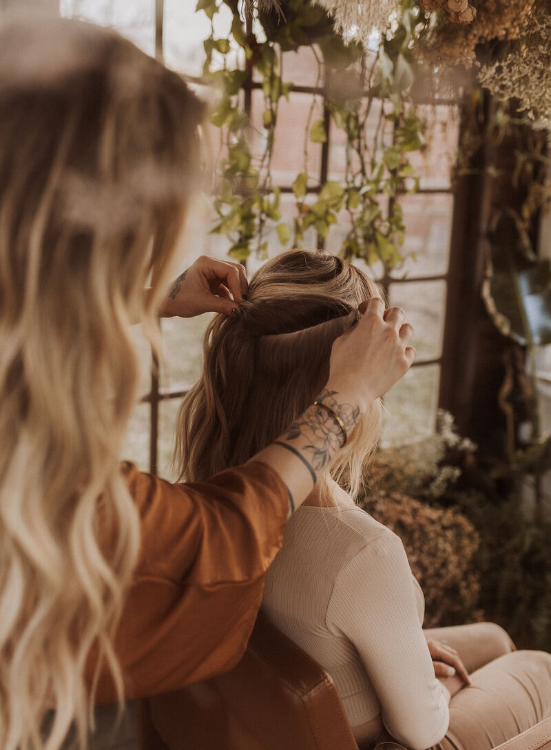 The hair extension expert is styling her client with long straight hair. Both women are in front of a mirror and her hand is tattooed with a butterfly.