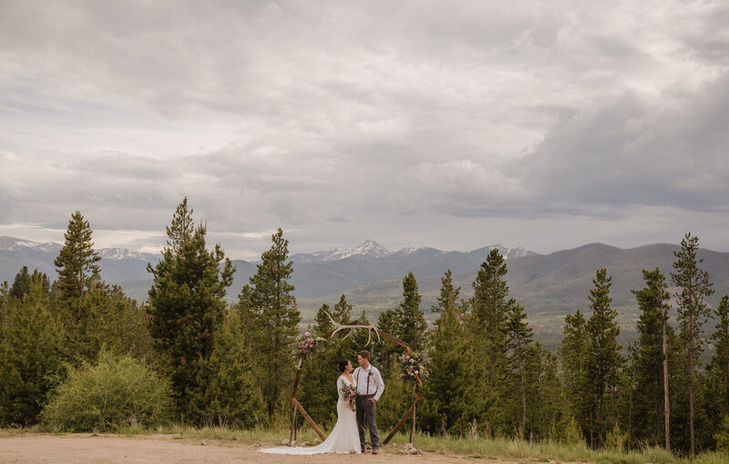 Bride and groom pose under their ceremony arch at their outdoor wedding in the mountains