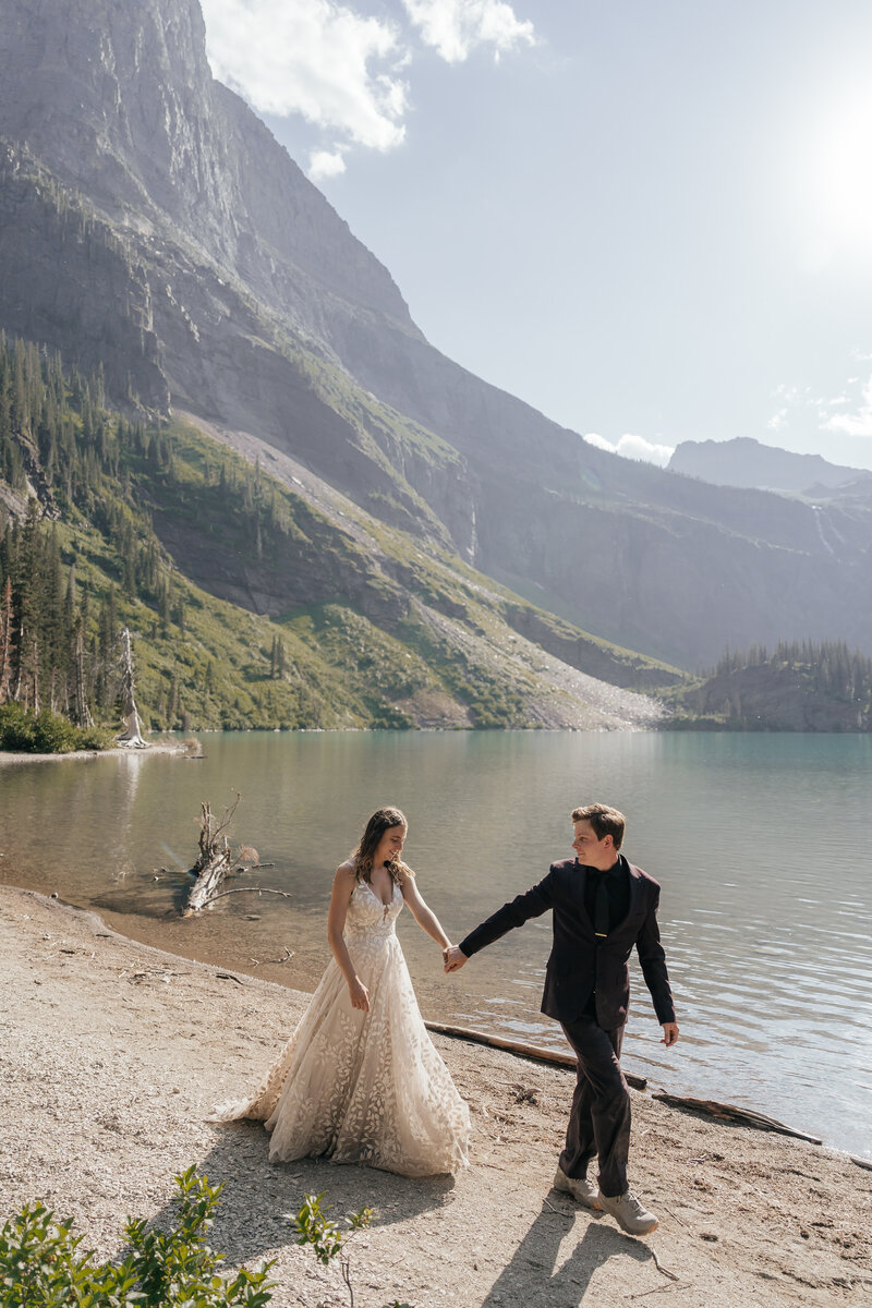 A couple holds hands and walks on the shore of an alpine lake in Glacier National Park.