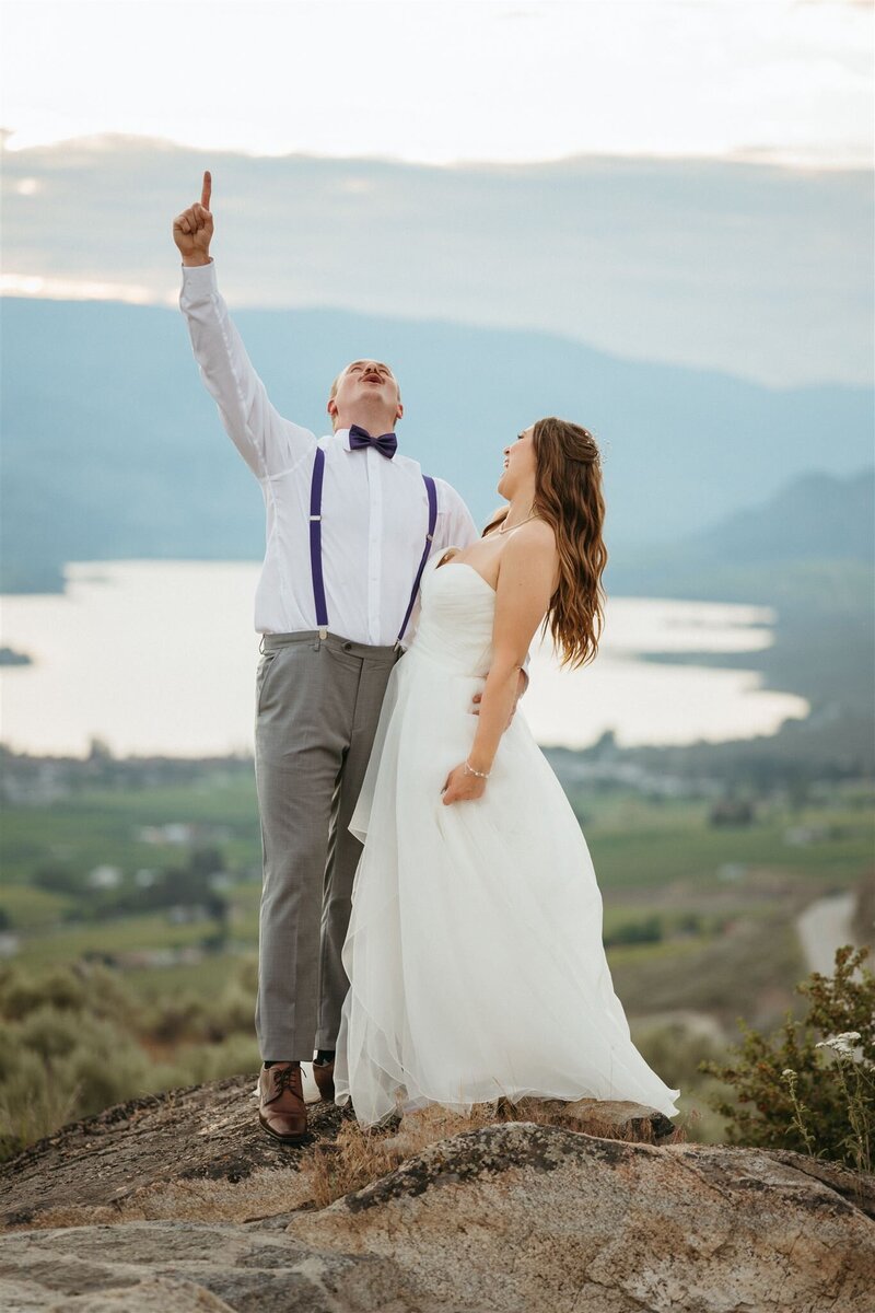 a couple on top of a mountain celebrating their wedding with joy and happiness- the bride is looking at the groom, wearing a white long wedding dress, the groom is looking up cheering wearing a white shirt with purple suspenders and bow tie, with brown shoes and gray pants