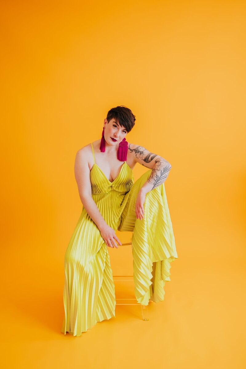 Woman with very short brown hair sits in a yellow chair wearing a bright yellow jumpsuit and long, red, fringe earrings while posing in front of yellow-orange backdrop.