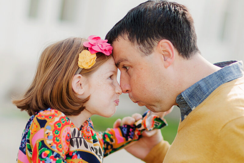 Girl with red hair and flower headband and Dad are nose-to-nose making a silly face.