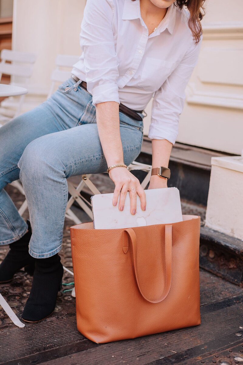 Madison reaching down to place her marble-patterned MacBook into a cognac leather tote, wearing jeans and a white blouse.