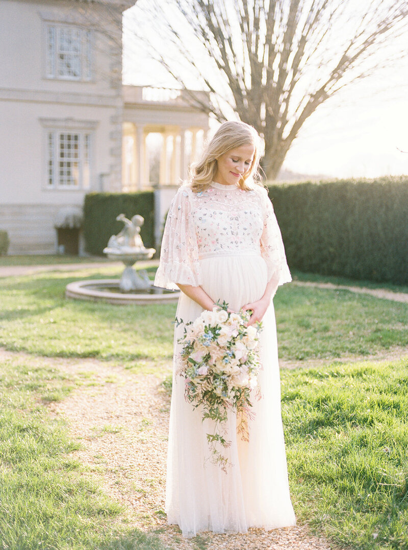Pregnant mother with blonde hair stands in Needle & Thread dress holding bouquet during her Maryland maternity session.