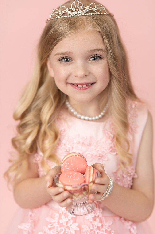 Young girl with long blonde hair wearing a sparkling tiara and pearl jewelry, smiling and holding pink macarons. The background is a soft pink tone.