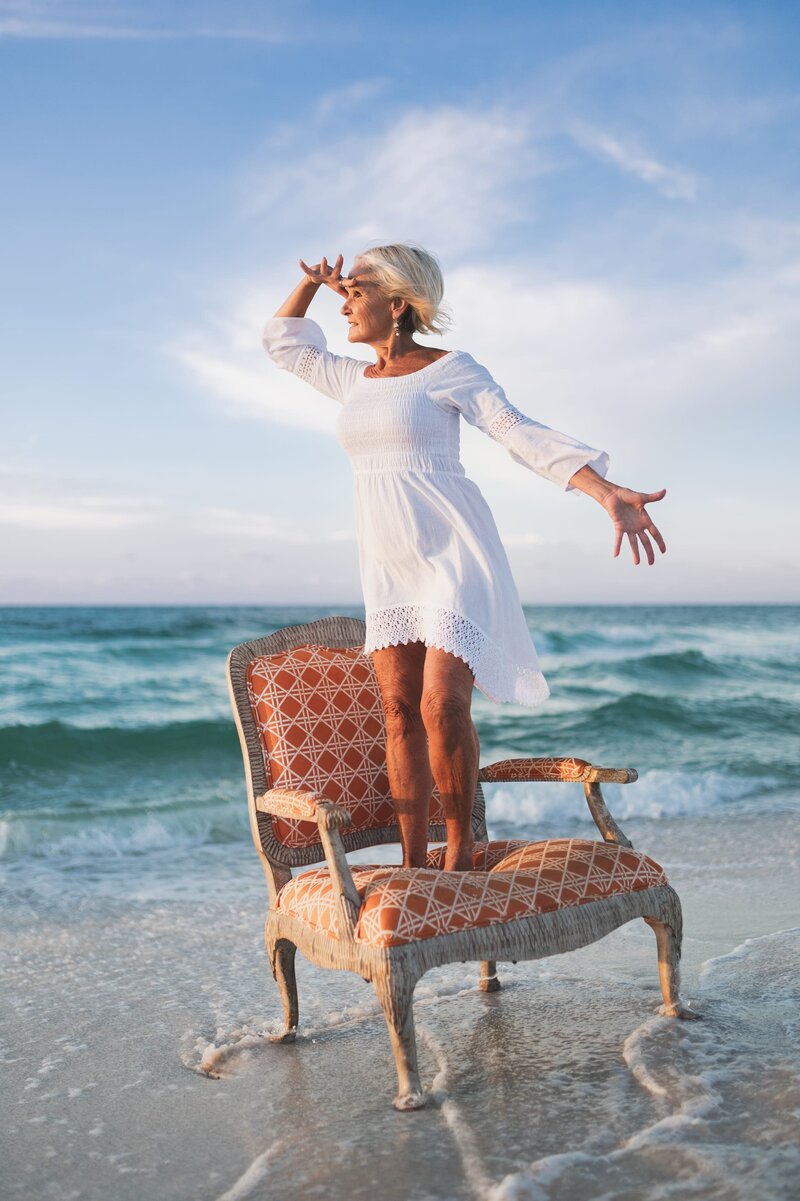 Older woman with white hair wearing a white long-sleeve dress stands on an antique chair on a Florida beach at sunset looking out into the distance.