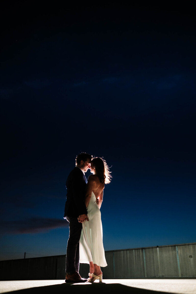 A couple stands close together under a deep blue night sky, illuminated by dramatic backlighting that highlights their embrace. The bride’s elegant white dress flows as they share a quiet, intimate moment, capturing the artistry and romance of Alabama wedding photography.