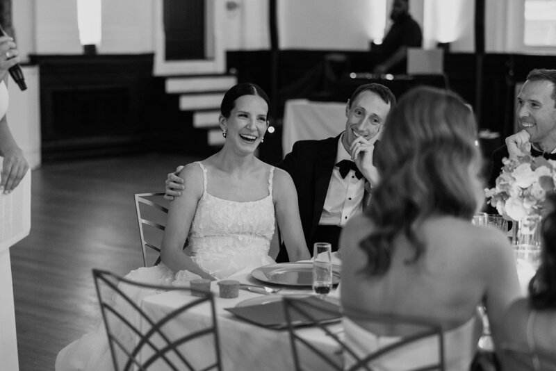 Black and white image of bride and groom smiling listening to toasts at their wedding reception in St. Louis
