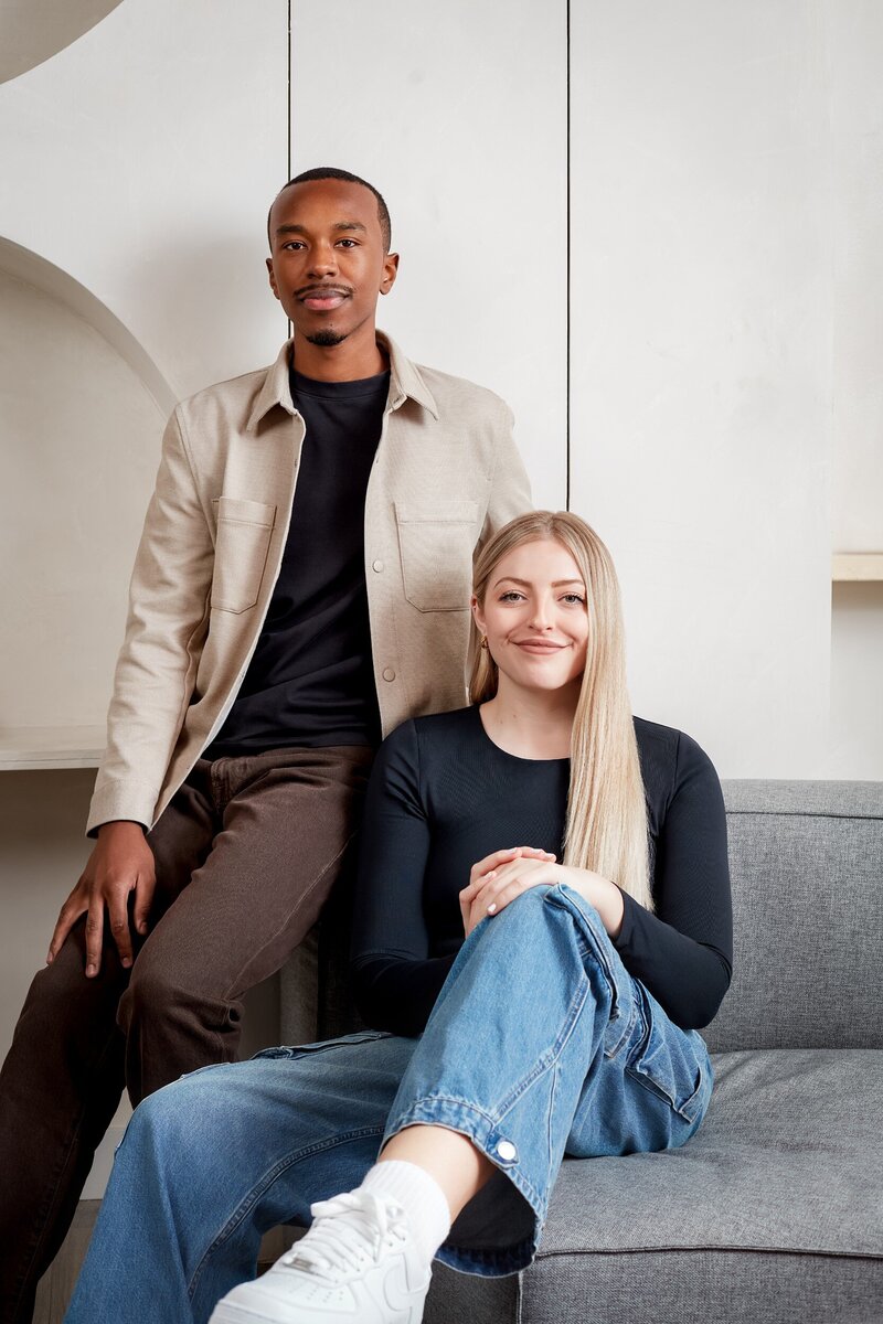 Steph and Dennis, Dennis is a young black man with short hair, and Steph is a young white woman with blonde hair. In this photo Steph is sitting on a grey sofa, and Dennis is sitting on the arm. They are wearing business casual clothes.