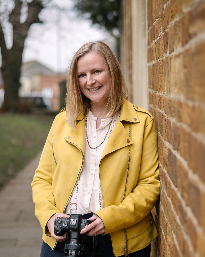 A headshot of Claire O'Brien Photography in a yellow jacket leaning on a wall
