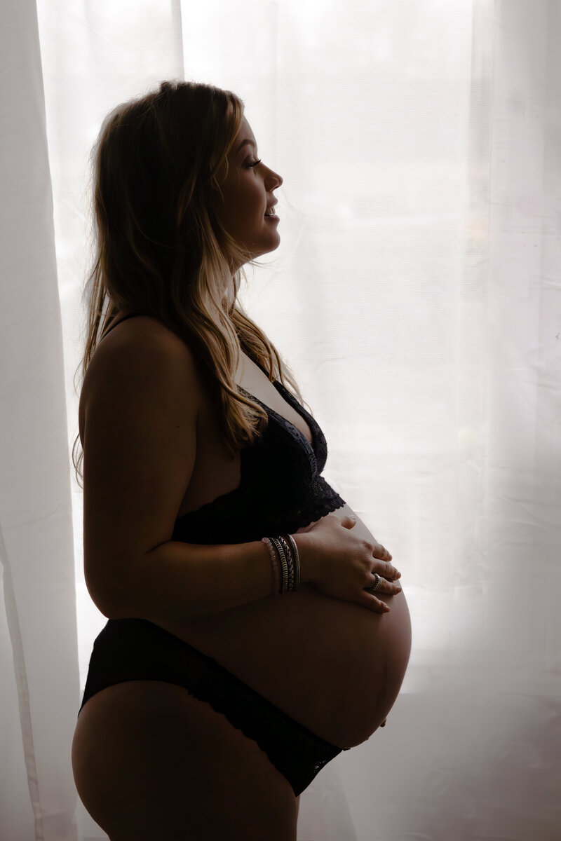 Zwangere vrouw in een zwarte beha en ondergoed, poseert met een serene uitstraling en een hand op haar buik.