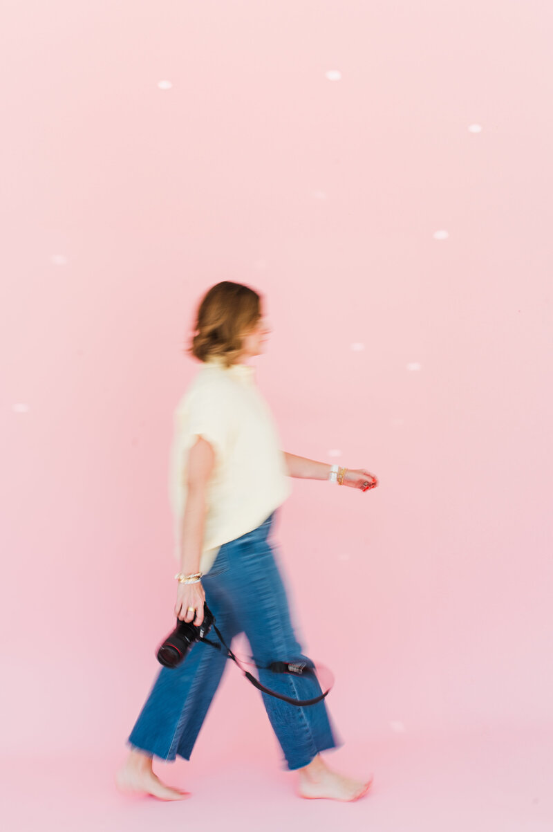 woman walking with camera in hand in front of a pink backdrop
