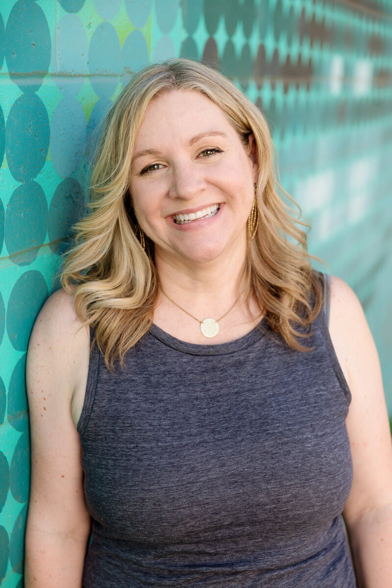 Calgary brand photography headshot of Lisa Sierra, a blond woman smiling, leaning against a colourful blue graffiti wall.