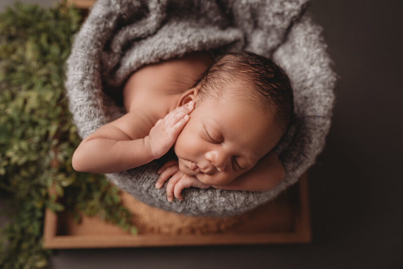 Newborn baby boy posed in wicker basket with chin on hand and other hand on cheek