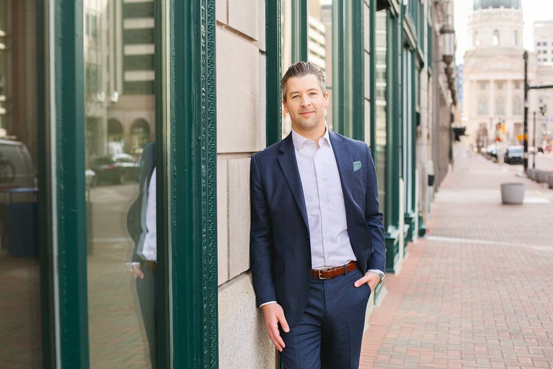 A-professional-man-in-a-navy-suit-smiling-confidently-during-a-headshot-session-in-downtown-Indianapolis-with-the-Indiana-Statehouse-in-the-background-on-the-circle-downtown-indy